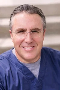 Dr. Christopher M. Sicher, D.M.D., smiling, in blue scrubs.